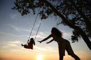silhouette of woman pushing child on a swing in a tree at sunset